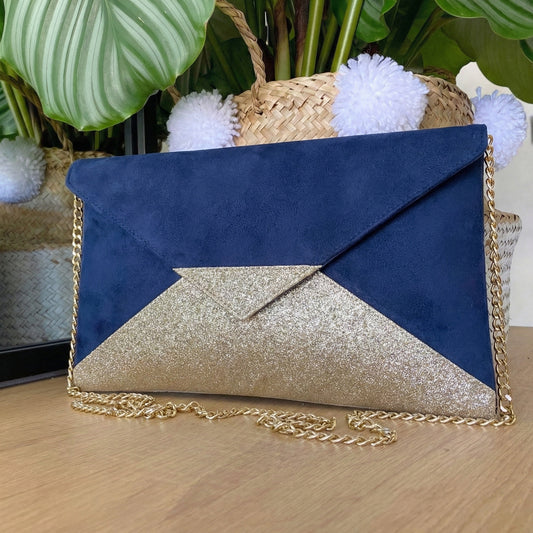 Pochette enveloppe avec chaîne dorée bi-matière suédine bleu marine et tissu pailleté or posée sur une surface bois