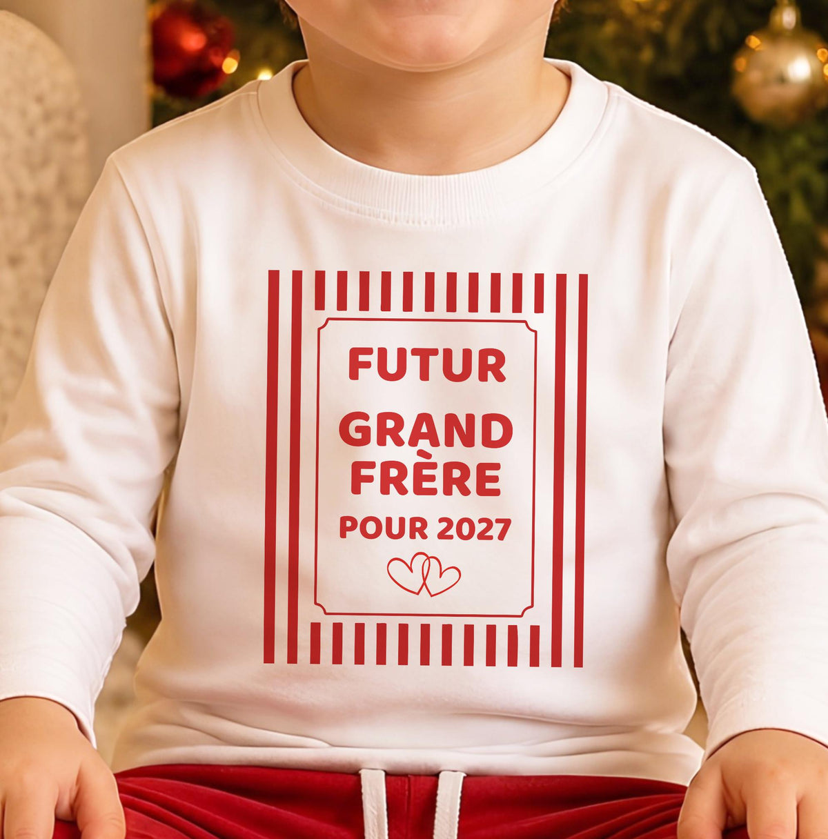 Gros plan du marquage rouge sur le t-shirt blanc : « Futur grand frère pour 2027 » dans un cadre à rayures façon sucre d'orge avec deux petits cœurs entrelacés