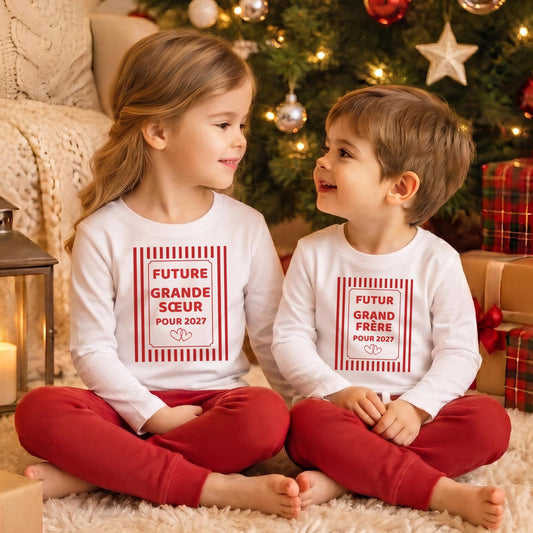 Frère et sœur assis côte à côte devant le sapin de Noël, portant les t-shirts assortis « Future grande sœur pour 2027 » et « Futur grand frère pour 2027 » à cadre rayé rouge, annonce de grossesse en famille