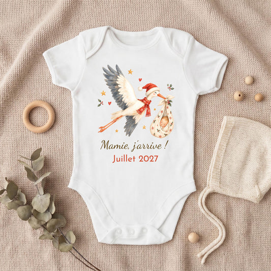 Body annonce grossesse cigogne de Noël avec le message Mamie, j'arrive ! Juillet 2027, posé sur un plaid beige avec bonnet bébé et anneau en bois