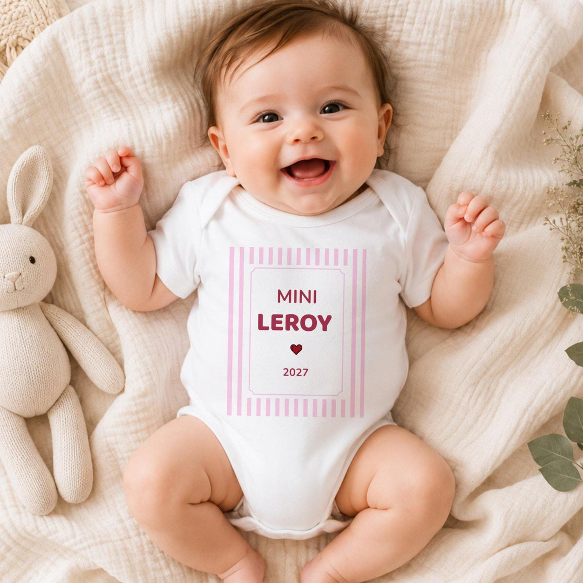 Bébé fille souriante portant un body blanc personnalisé « MINI LEROY 2027 » en framboise dans un cadre à rayures roses, allongée sur une couverture en gaze crème à côté d'un lapin en tricot