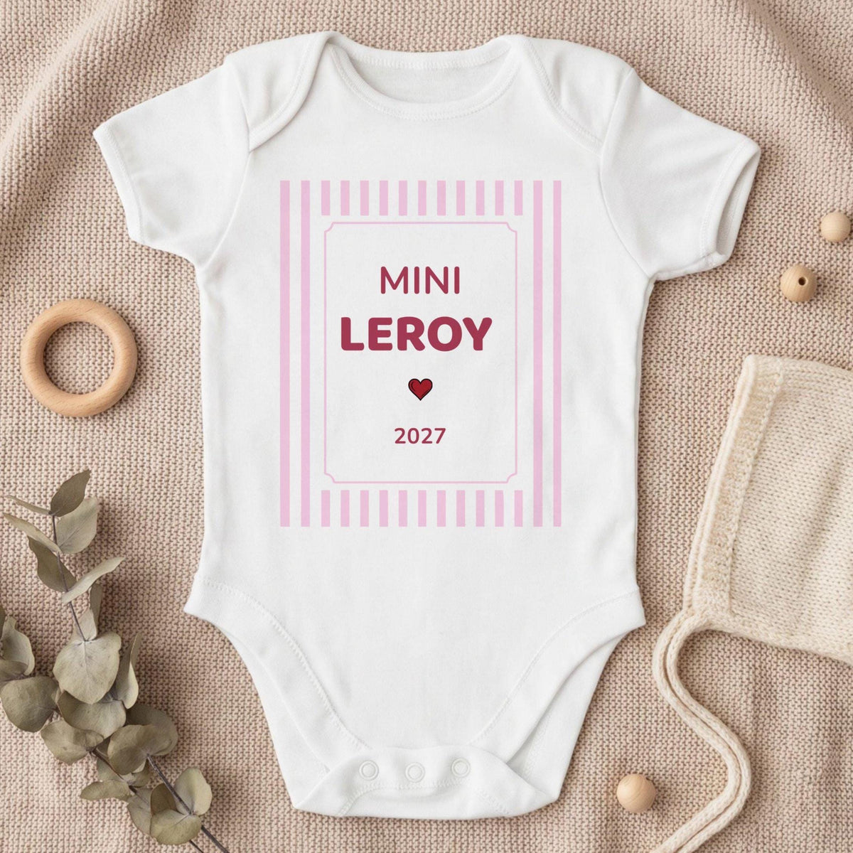 Body bébé fille blanc personnalisé « MINI LEROY 2027 » en framboise avec un petit cœur rouge, dans un cadre à rayures roses, posé à plat sur un plaid en tricot beige avec eucalyptus et béguin