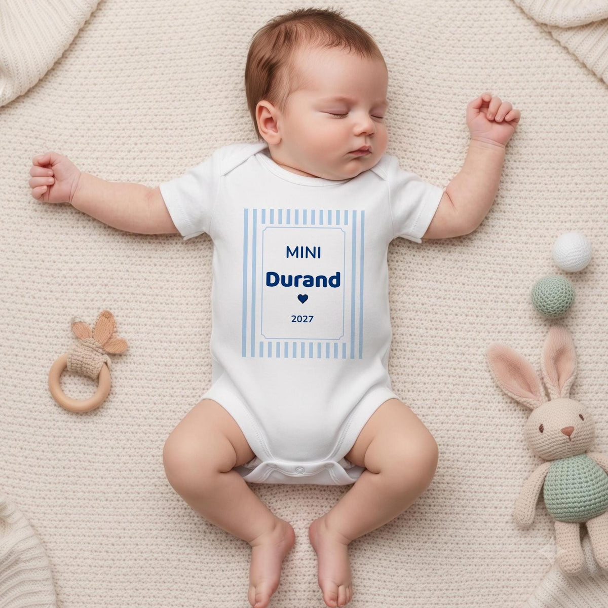 Nouveau-né endormi portant un body blanc personnalisé « MINI Durand 2027 » en bleu marine dans un cadre à rayures bleu ciel, allongé sur un plaid crème avec un lapin en crochet et un hochet en bois