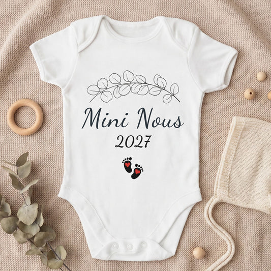 Body bébé blanc personnalisé « Mini Nous 2027 » avec branche d'eucalyptus et petites empreintes de pieds en forme de cœur, posé sur un plaid en tricot beige