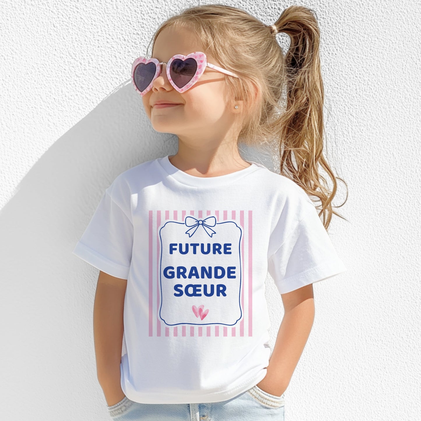 T-shirt enfant blanc « Future grande sœur » porté par une petite fille, motif rayures roses et nœud bleu marine