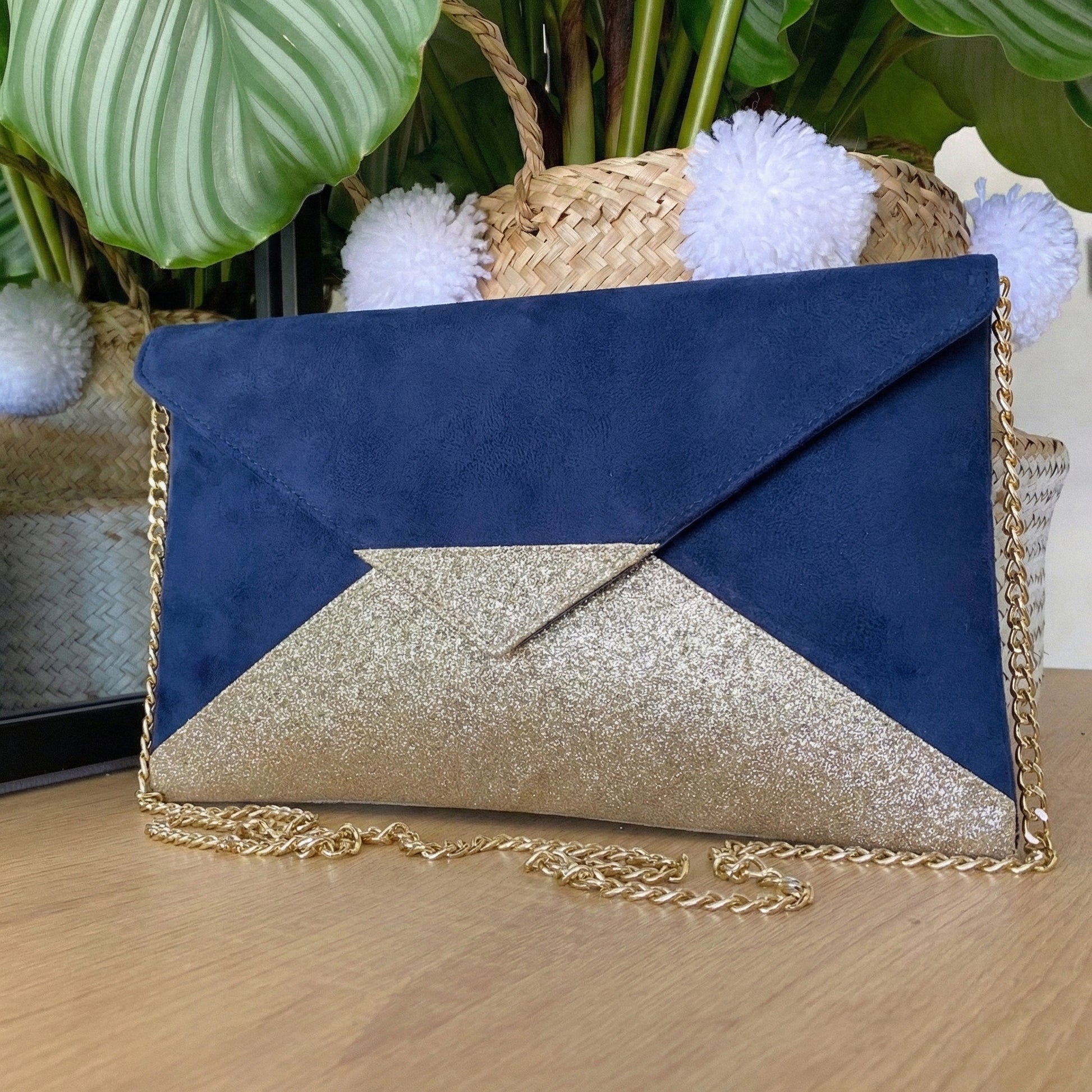 Pochette enveloppe  avec chaîne dorée bi-matière suédine bleu marine et tissu pailleté or posée sur une surface bois