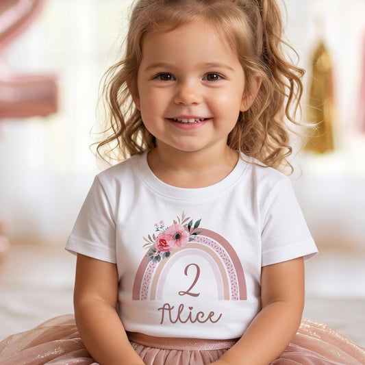Petite fille souriante portant un t-shirt personnalisé pour anniversaire, motif arc-en-ciel bohème fleuri rose avec le chiffre 2 et le prénom Alice floqués en flex rose gold