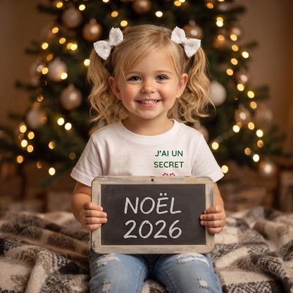 Petite fille tenant un panneau « Noël 2026 », tee-shirt annonce grossesse « J'ai un secret »