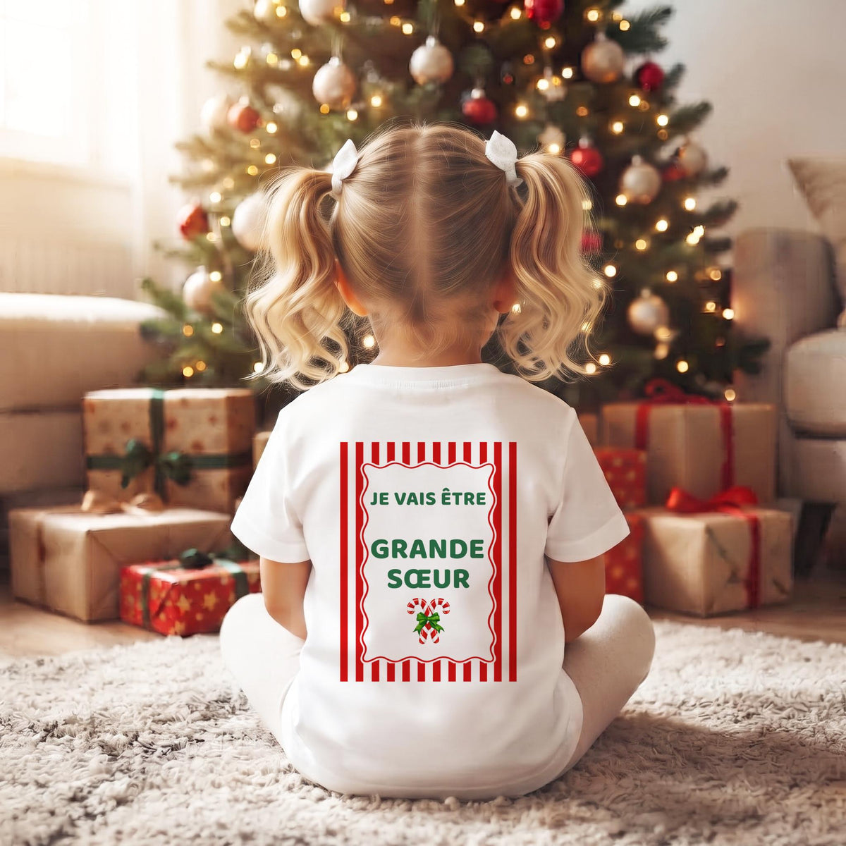 Petite fille assise de dos devant le sapin de Noël, tee-shirt « Je vais être Grande Sœur » à rayures rouges