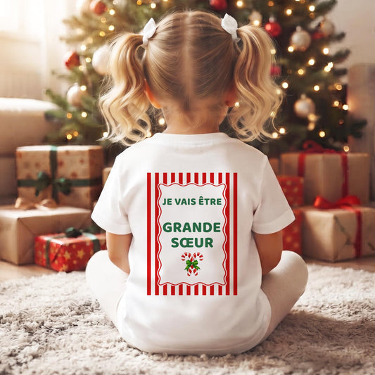 Petite fille assise de dos devant le sapin de Noël, tee-shirt « Je vais être Grande Sœur » à rayures rouges