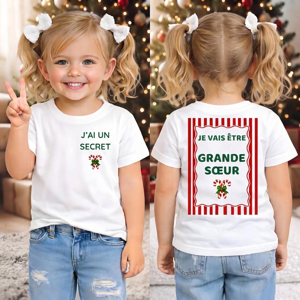 Deux fillettes aux couettes blondes portant le tee-shirt annonce grossesse de Noël, avant « J'ai un secret » et dos « Je vais être Grande Sœur », devant un sapin décoré