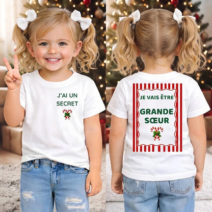 Deux fillettes aux couettes blondes portant le tee-shirt annonce grossesse de Noël, avant « J'ai un secret » et dos « Je vais être Grande Sœur », devant un sapin décoré