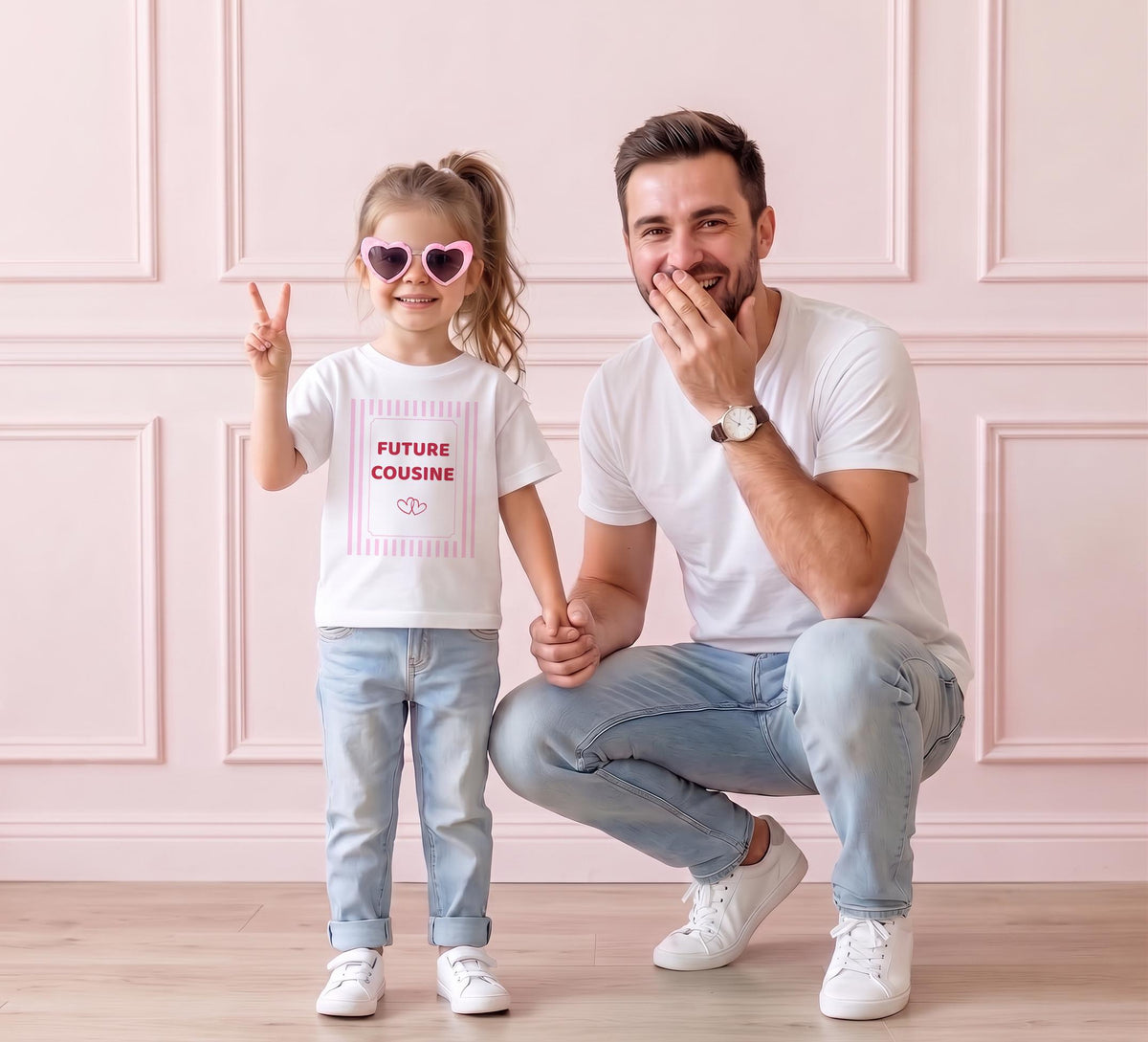 Petite fille et son papa souriants, elle porte le tee-shirt « FUTURE COUSINE » à rayures roses en faisant un signe de victoire, devant un mur rose pastel, pour une annonce de grossesse en famille.