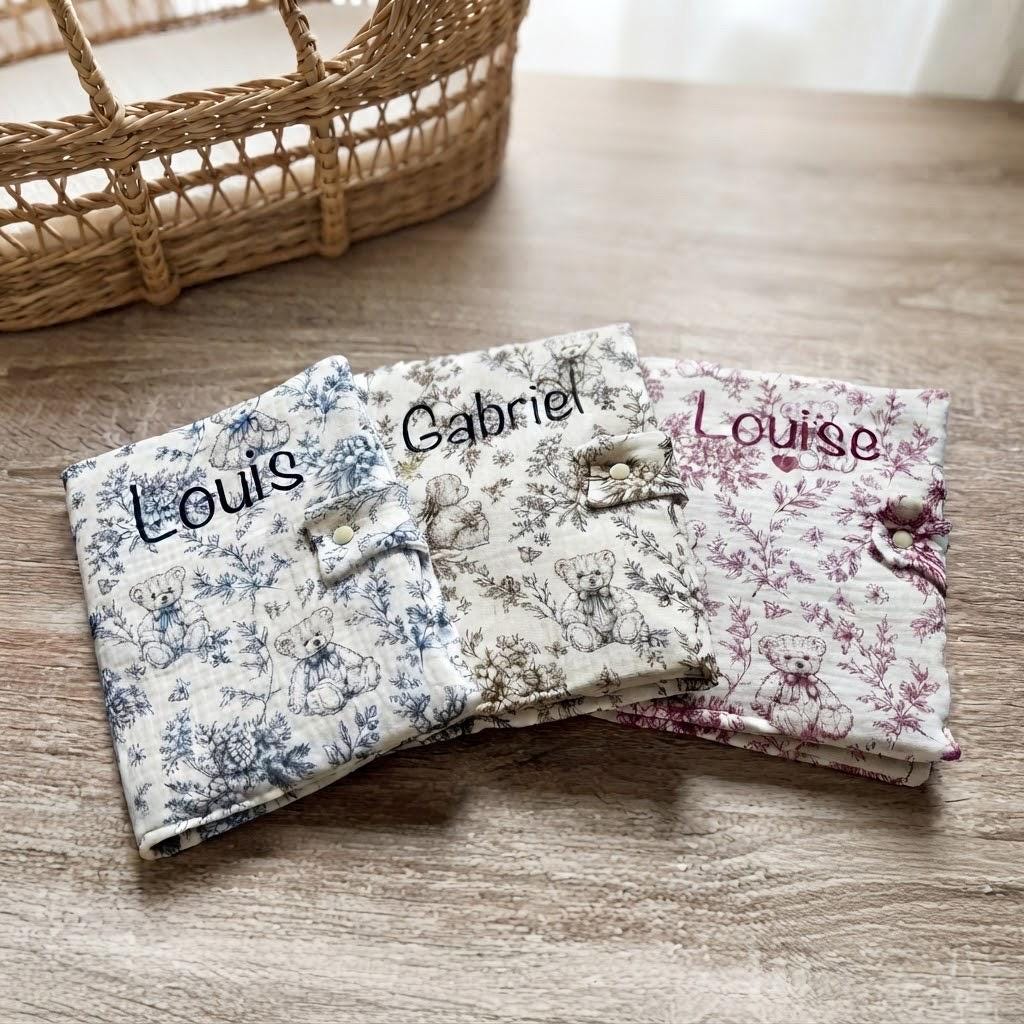 Trois protège carnets de santé en gaze de coton motif toile de Jouy oursons, coloris bleu, marron et bordeaux, personnalisés aux prénoms Louis, Gabriel et Louise, avec un cœur sous Louise