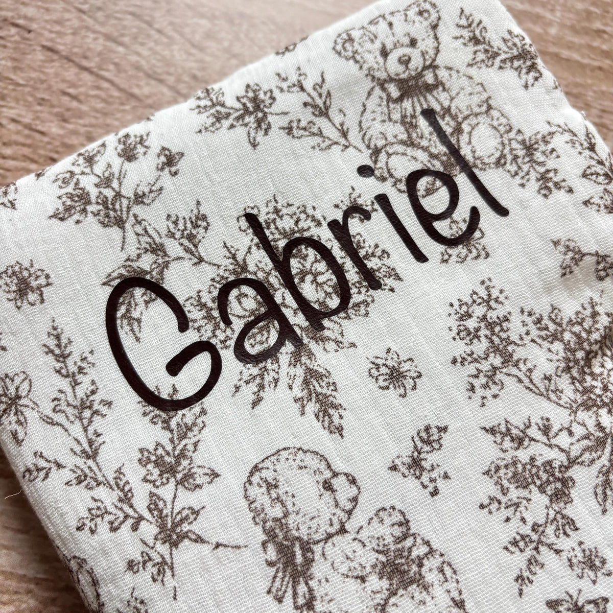 Gros plan sur le prénom Gabriel floqué en marron foncé sur la gaze de coton gaufrée motif toile de Jouy oursons du protège carnet de santé