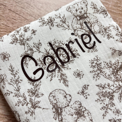 Gros plan sur le prénom Gabriel floqué en marron foncé sur la gaze de coton gaufrée motif toile de Jouy oursons du protège carnet de santé