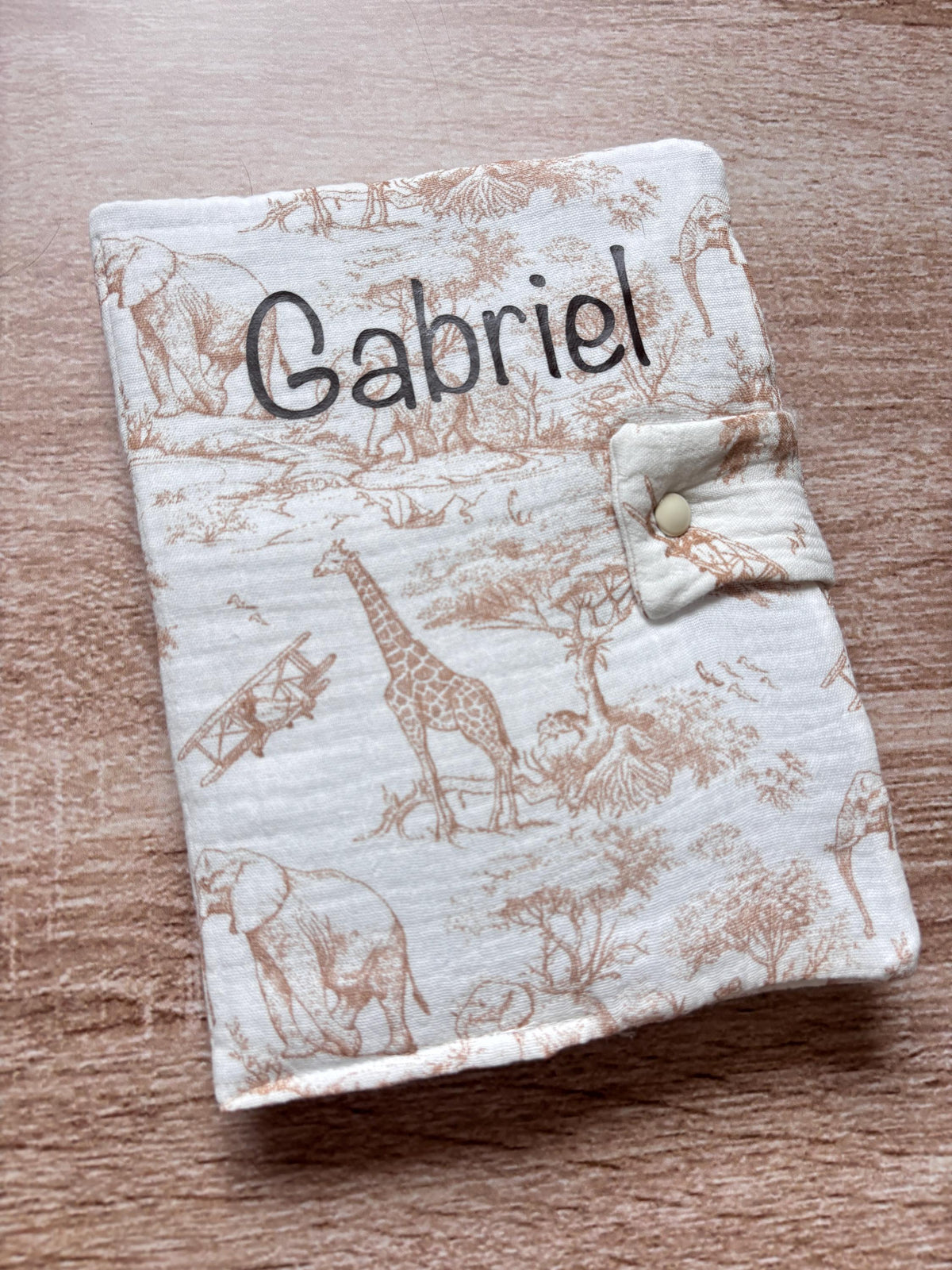 Protège carnet de santé beige en gaze de coton motif toile de Jouy Safari avec girafe et éléphants, personnalisé au prénom Gabriel en flocage marron chocolat, fermeture par patte à bouton pression