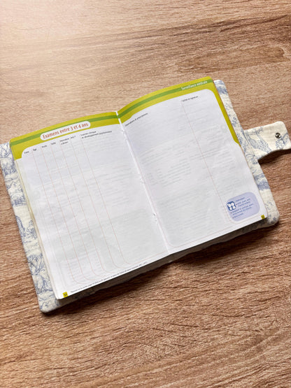 Protège carnet de santé bleu en gaze de coton motif toile de Jouy Safari ouvert, avec le carnet de santé glissé dans les rabats intérieurs blancs et la patte à bouton pression