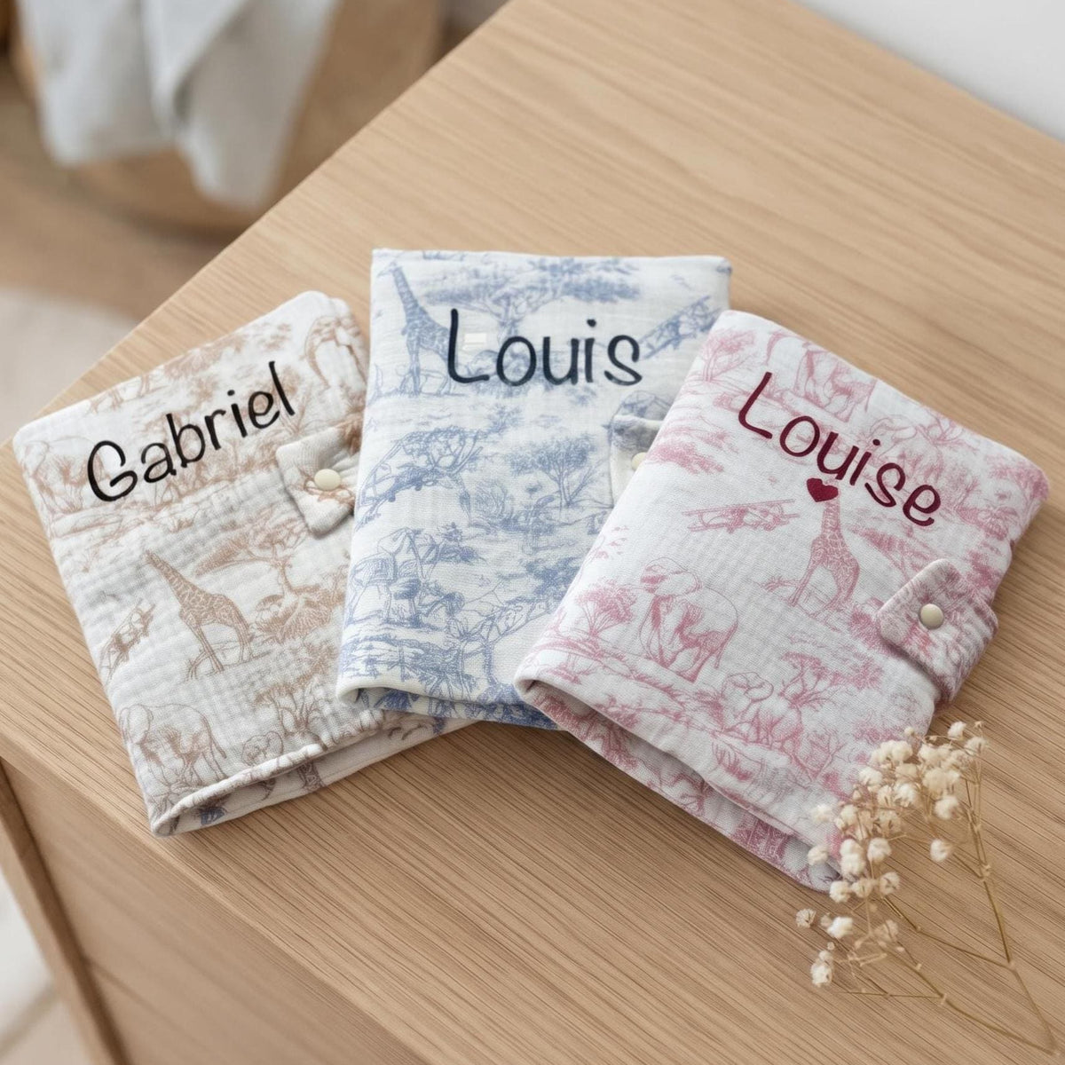 Trois protège carnets de santé en gaze de coton motif toile de Jouy Safari, coloris beige, bleu et rose, personnalisés aux prénoms Gabriel, Louis et Louise, avec un cœur sous Louise