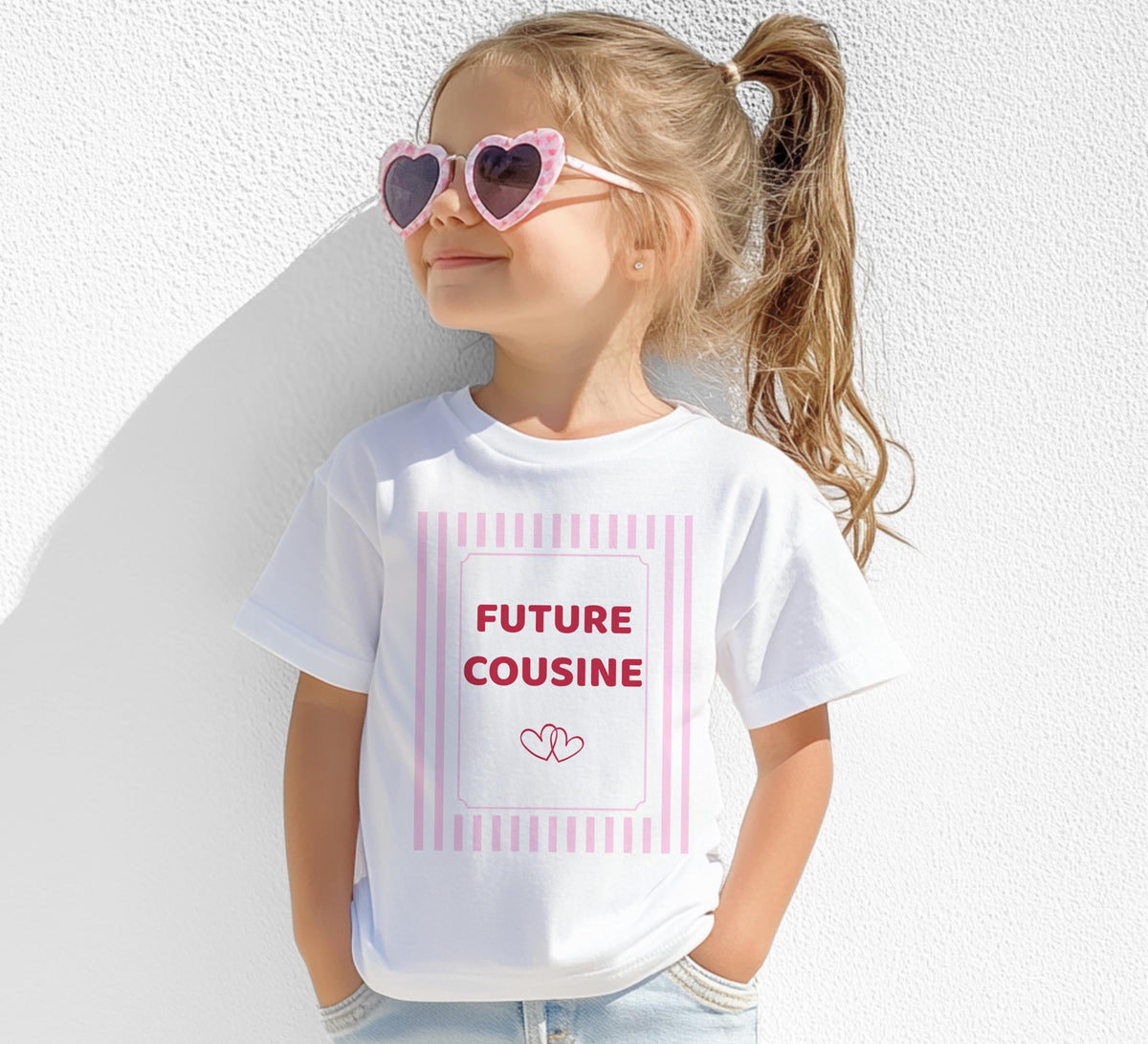 Petite fille portant des lunettes de soleil roses en forme de cœur et un tee-shirt blanc floqué « FUTURE COUSINE » dans un cadre à rayures roses avec deux petits cœurs, photographiée de face devant un mur blanc.