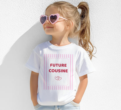 Petite fille portant des lunettes de soleil roses en forme de cœur et un tee-shirt blanc floqué « FUTURE COUSINE » dans un cadre à rayures roses avec deux petits cœurs, photographiée de face devant un mur blanc.