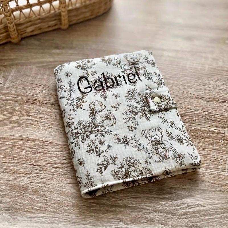 Protège carnet de santé marron en gaze de coton motif toile de Jouy oursons et fleurs, personnalisé au prénom Gabriel en flocage marron foncé, avec patte à bouton pression