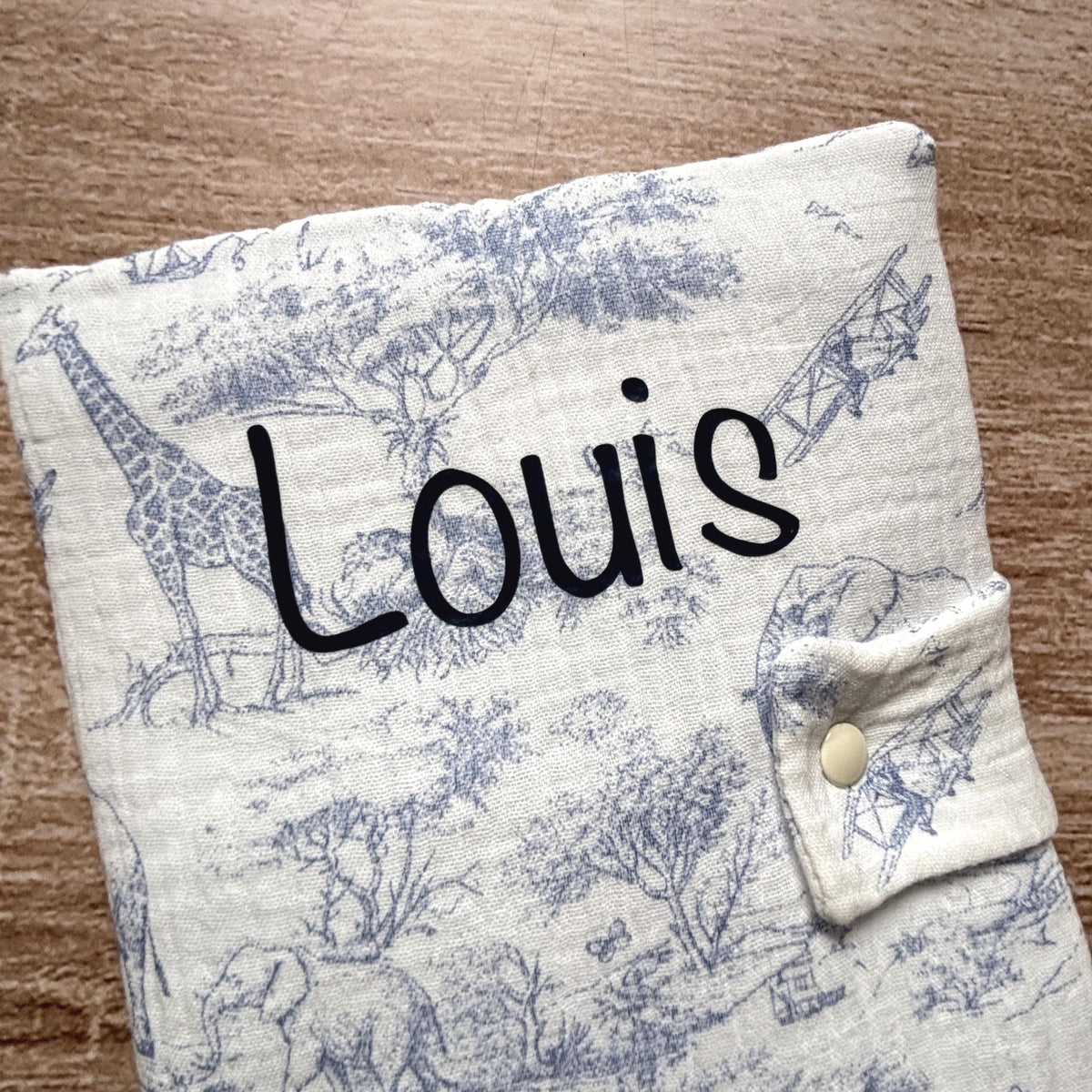 Gros plan sur le prénom Louis floqué en bleu marine sur la gaze de coton gaufrée motif toile de Jouy Safari du protège carnet de santé