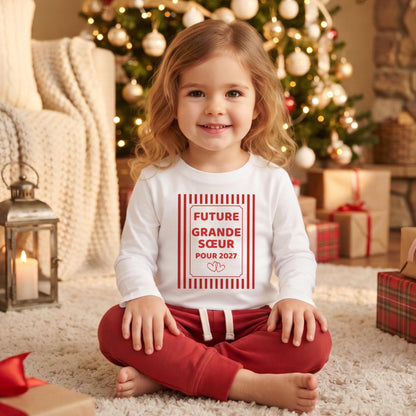 Petite fille assise devant le sapin de Noël portant un t-shirt blanc manches longues « Future grande sœur pour 2027 » dans un cadre à rayures rouges avec deux cœurs, idée d'annonce de grossesse à Noël
