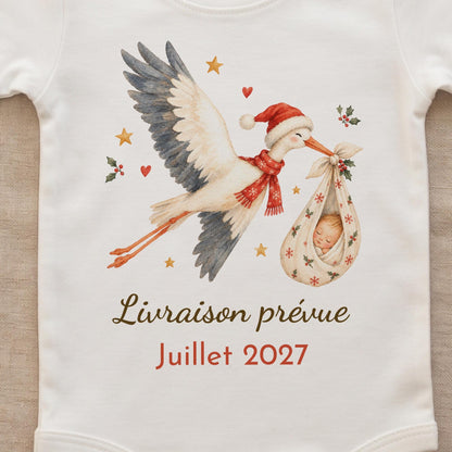 Gros plan sur l'impression du body : cigogne de Noël aquarelle avec bonnet et écharpe rouge, bébé endormi dans un baluchon à flocons et houx, texte Livraison prévue Juillet 2027