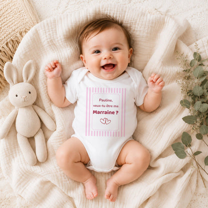 Bébé souriant portant un body blanc personnalisé « Pauline, veux-tu être ma Marraine ? » dans un cadre à rayures roses
