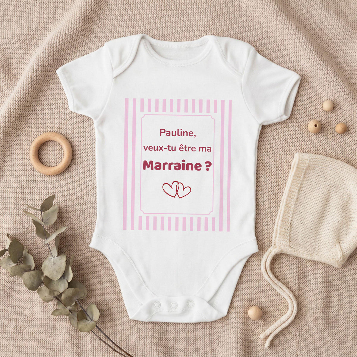 Body bébé blanc personnalisé avec cadre à rayures roses et texte framboise « Pauline, veux-tu être ma Marraine ? » et deux cœurs entrelacés, posé à plat sur un plaid en tricot beige