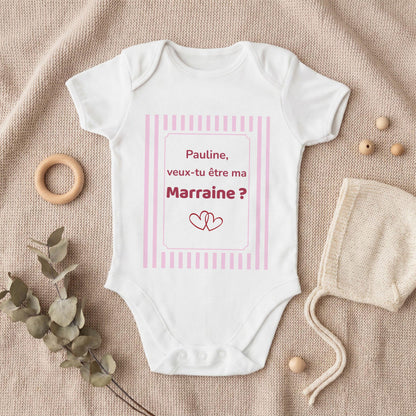 Body bébé blanc personnalisé avec cadre à rayures roses et texte framboise « Pauline, veux-tu être ma Marraine ? » et deux cœurs entrelacés, posé à plat sur un plaid en tricot beige