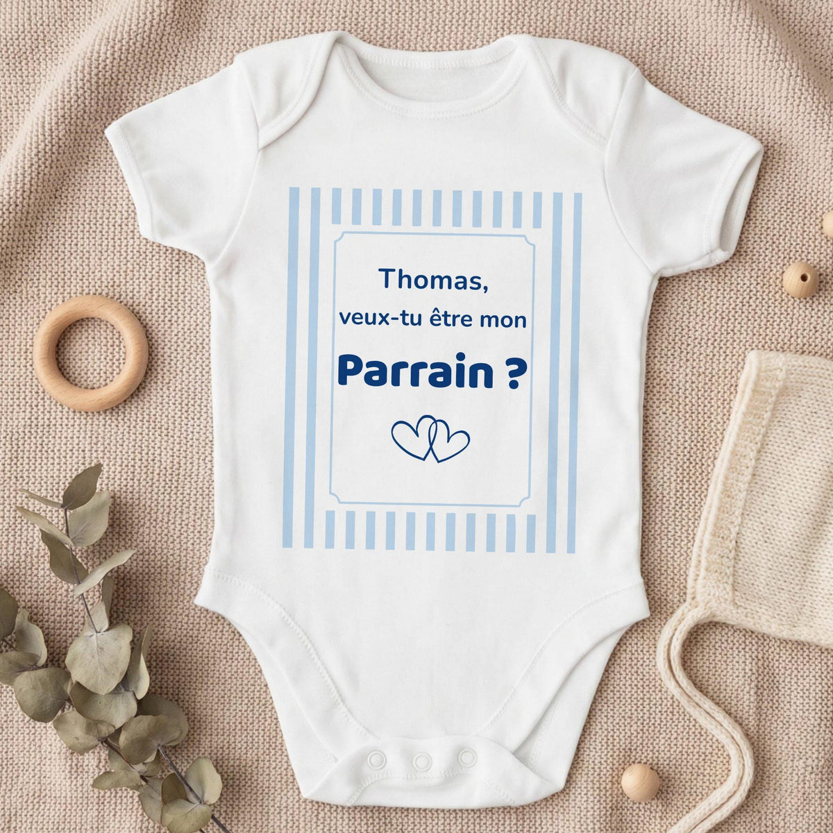 Body bébé blanc personnalisé avec cadre à rayures bleu ciel et texte bleu marine « Thomas, veux-tu être mon Parrain ? » et deux cœurs entrelacés, posé à plat sur un plaid en tricot beige