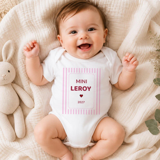 Bébé fille souriante portant un body blanc personnalisé « MINI LEROY 2027 » en framboise dans un cadre à rayures roses, allongée sur une couverture en gaze crème à côté d'un lapin en tricot