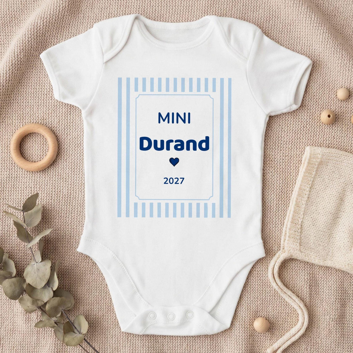 Body bébé garçon blanc personnalisé « MINI Durand 2027 » en bleu marine avec un petit cœur, dans un cadre à rayures bleu ciel, posé à plat sur un plaid en tricot beige avec eucalyptus et béguin