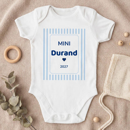 Body bébé garçon blanc personnalisé « MINI Durand 2027 » en bleu marine avec un petit cœur, dans un cadre à rayures bleu ciel, posé à plat sur un plaid en tricot beige avec eucalyptus et béguin