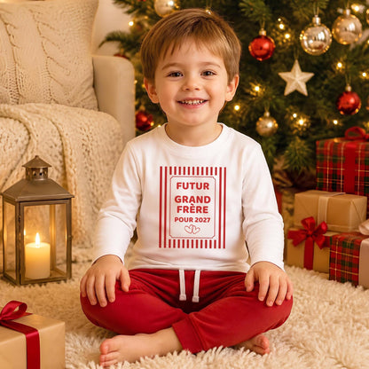 Petit garçon souriant au pied du sapin de Noël portant un t-shirt blanc manches longues « Futur grand frère pour 2027 » dans un cadre à rayures rouges avec deux cœurs, révélation bébé à Noël