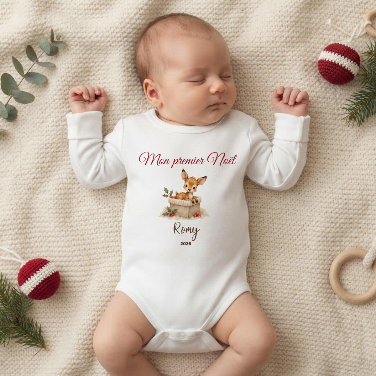 Bébé endormi portant le body Mon premier Noël personnalisé au prénom, motif faon et houx, sur un plaid crème avec boules de Noël rouges