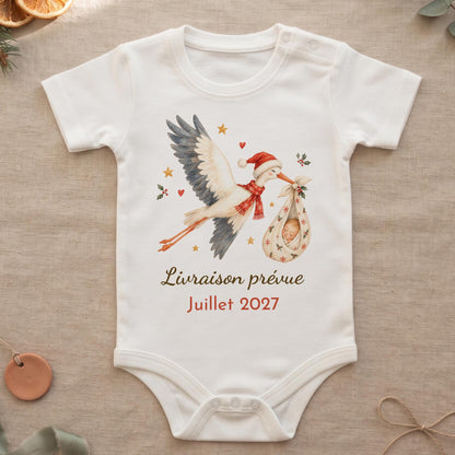 Body bébé blanc manches courtes personnalisé pour annonce de grossesse à Noël : cigogne aquarelle en bonnet de Noël portant bébé dans un baluchon, texte Livraison prévue Juillet 2027, posé sur du lin avec oranges séchées et eucalyptus
