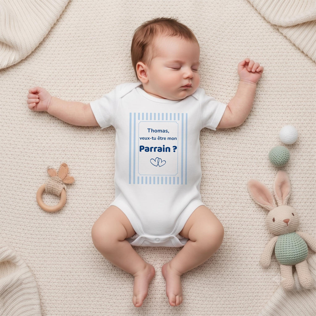 Nouveau-né endormi portant un body blanc personnalisé « Thomas, veux-tu être mon Parrain ? » dans un cadre à rayures bleu ciel