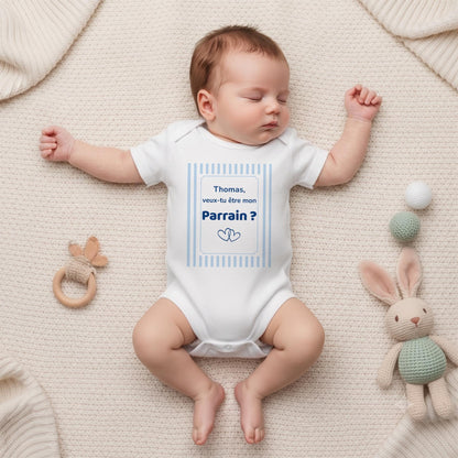 Nouveau-né endormi portant un body blanc personnalisé « Thomas, veux-tu être mon Parrain ? » dans un cadre à rayures bleu ciel