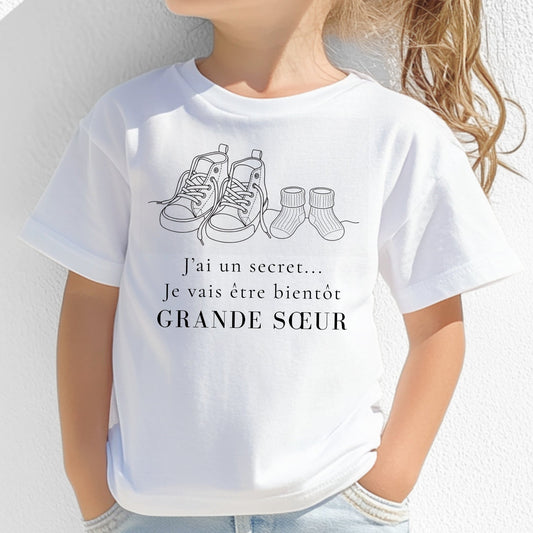 T-shirt enfant blanc « J'ai un secret, je vais être bientôt grande sœur » avec illustration line art de baskets et chaussettes de bébé