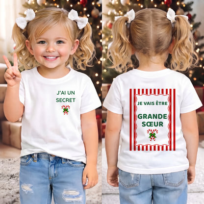 Tee-shirt enfant annonce grossesse de Noël « J'ai un secret » / « Je vais être Grande Sœur », porté devant un sapin