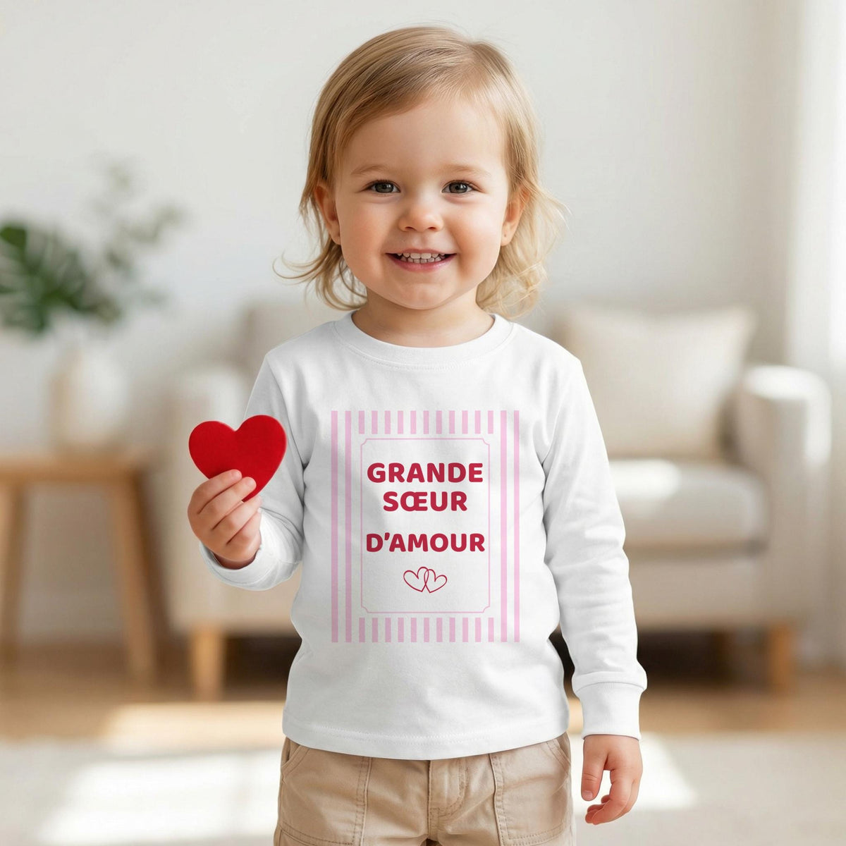 Petite fille souriante tenant un cœur rouge, vêtue d'un t-shirt blanc manches longues Grande sœur d'amour, cadre à rayures roses et deux cœurs
